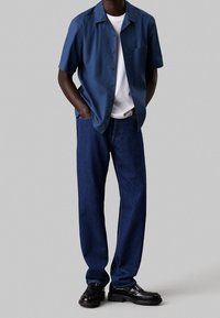 Mies, jolla on yllään tummansininen lyhythihainen paita valkoisen T-paidan päällä, siniset farkut ja mustat nahkakengät, seisoo käsivartensa taskuissa.