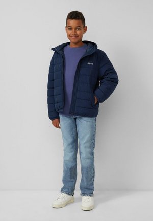 Marineblaue Pufferjacke mit Reißverschluss und Kapuze, ausgestattet mit einer strukturierten Oberfläche. Trägt einen lila Pullover, kombiniert mit hellblauen Jeans und weißen Turnschuhen.
