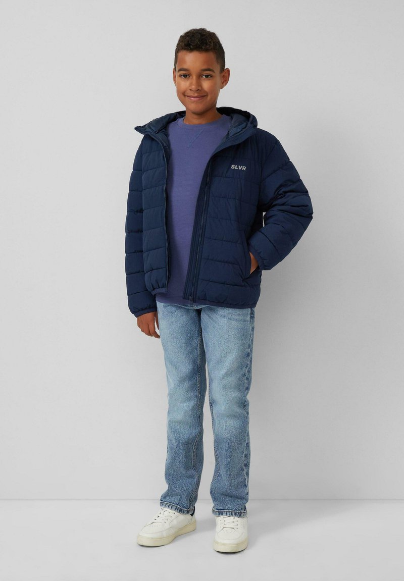 Marineblaue Pufferjacke mit Reißverschluss und Kapuze, ausgestattet mit einer strukturierten Oberfläche. Trägt einen lila Pullover, kombiniert mit hellblauen Jeans und weißen Turnschuhen.