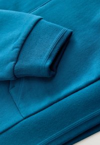 Felpa in teal con polsini e bordo a costine, realizzata in tessuto morbido con una texture liscia. Include un dettaglio a tasca a marsupio.