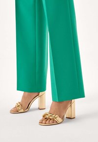 Guld-sandaler med blokhæl og bred rem med kædedetalje, kombineret med livlige grønne bukser med vide ben.