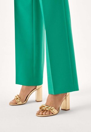 Guld-sandaler med blokhæl og bred rem med kædedetalje, kombineret med livlige grønne bukser med vide ben.