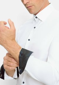 Weißes Hemd mit Knopfleiste aus strukturiertem Stoff. Dunkle, gemusterte Manschetten stehen im Kontrast zum weißen Körper und besitzen blaue Knöpfe.