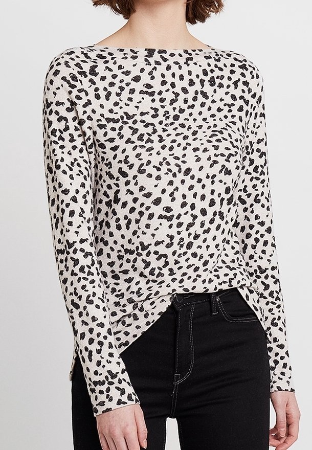 Femme portant un haut blanc à manches longues avec des taches noires irrégulières et un jean noir taille haute sur un fond uni.