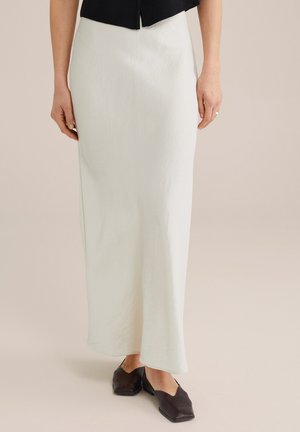 Jupe blanche longue jusqu'au sol avec une surface lisse et légèrement texturée. Associée à un haut noir court et des chaussures ouvertes brun foncé.