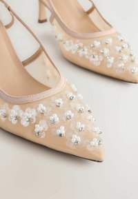 Pantofi cu toc bej, cu vârf ascuțit și finisaj lucios, decorați cu ornamente florale albe și accente mici argintii. Include toc subțire și detalii din plasă.