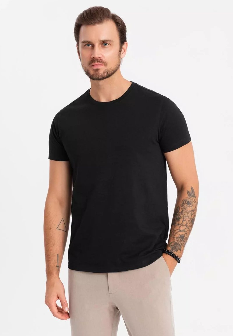 Czarny, krótki rękaw T-shirt wykonany z bawełny, z okrągłym dekoltem. Model ma tatuaże na ramieniu i nosi jasne spodnie.