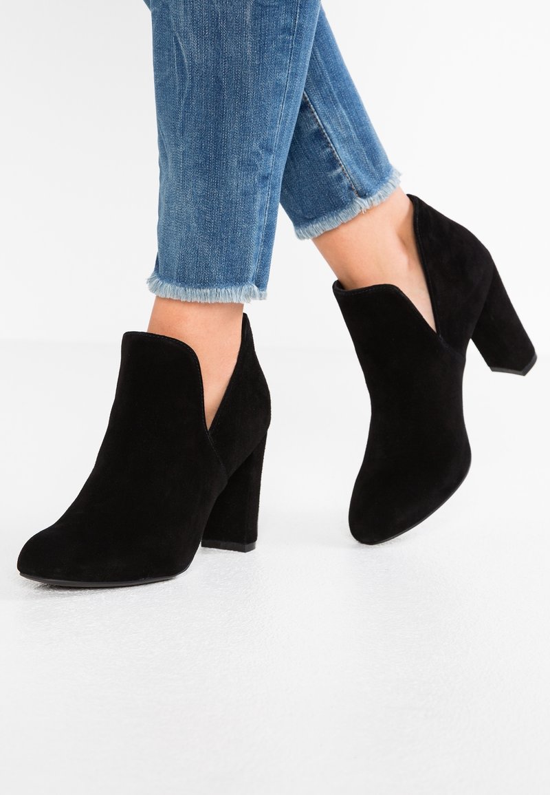 Botines de ante negros con tacón alto y cuadrado, que presentan un diseño en forma de V en los laterales y una textura suave. Combinados con vaqueros cortos.