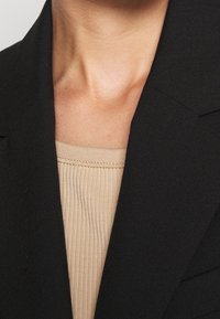 Svart kavaj med en strukturerad krage, kombinerad med en beige ribbad topp. Fokusera på tygtextur och färgkontrast.