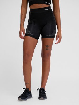 Osoba nosząca czarne sportowe spodenki Hummel oraz czarne trampki, prezentująca tatuaże na nogach i rękach na jednolitym szarym tle.