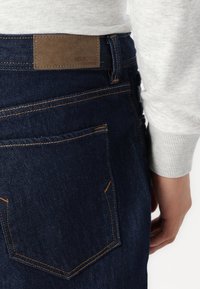 Mörkblå denim jeans med en läderetikett som läser "SELECTED HOMME," med orange sömmar och en slim fit-design.