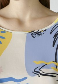Top a maniche corte multicolore con motivi astratti gialli, blu e neri su uno sfondo chiaro e scollatura a barchetta.