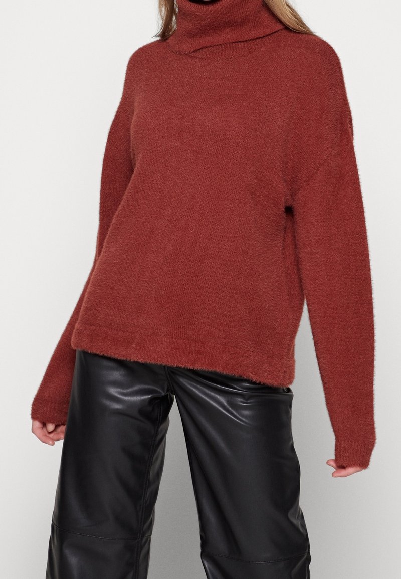 Chunčový rolák v červenohnědé barvě, vyrobený z huňatého materiálu, s vykrojenými rameny a uvolněným střihem, doplněný černými koženými kalhotami.