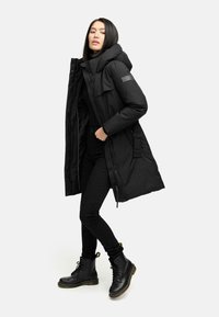 Sort pufferjakke med høj krave og fastgjort hætte, der har lynlås og trykknaplukning, parret med sorte skinny jeans og støvler.