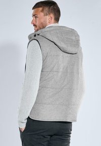 Mann trägt eine hellgraue gesteppte Weste mit Kapuze über einem weißen, gerippten Pullover und dunklen Hosen, schaut nach links und hat die Hände in den Taschen.