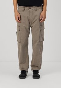 Beige cargo bukser laget av bomullsblend, med flere sidelommer og rett ben-design. Kombinert med svarte joggesko.