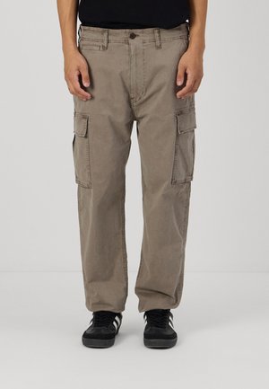 Pantalons cargo beiges en tissu mélangé coton, avec de multiples poches latérales et une coupe droite. Associés à des baskets noires.