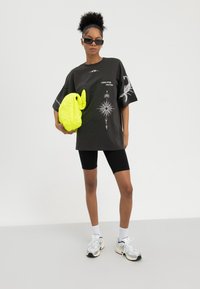 Frau mit lockigem Haar, die ein dunkles, grafisches, übergroßes T-Shirt, schwarze Radlerhosen, weiße Sneaker und Sonnenbrille trägt und eine leuchtend gelbe, gesteppte Tasche vor weißem Hintergrund hält.