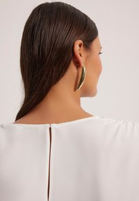 Biała bluzka z rozcięciem z tyłu, zestawiona z długimi, błyszczącymi złotymi kolczykami w kształcie hoop. Gładka faktura materiału i eleganckie włosy.
