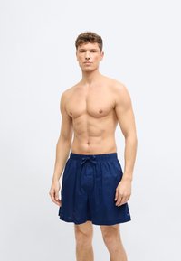 Άνδρας χωρίς μπλούζα με σγουρά μαλλιά, φοράει μπλε ναυτικό μαγιό με κορδόνι, στέκεται μπροστά σε απλό φωτεινό φόντο, κοιτώντας προς τα εμπρός.