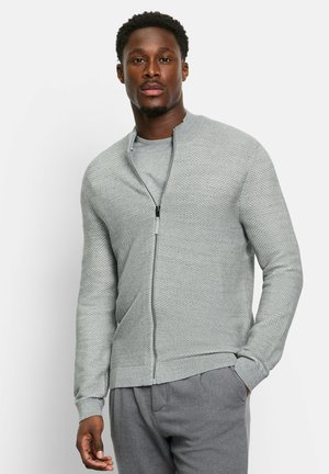 Hellgraue Zip-Up-Strickjacke mit einem strukturierten Design und Rippbündchen. Über einem passenden grauen T-Shirt getragen, kombiniert mit grauen Hosen.