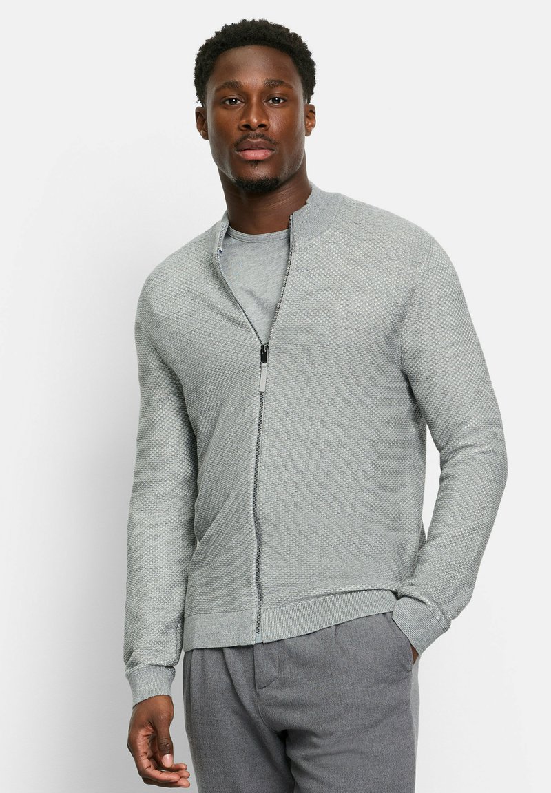 Lichtgrijze ritsvest met een textuurpatroon en ribgebreide manchetten. Gedragen over een bijpassend grijs T-shirt, gecombineerd met grijze broek.
