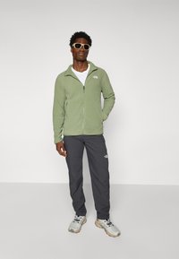 Veste en polaire vert clair avec une fermeture éclair à l'avant, assortie à un pantalon gris foncé et des baskets grises. Les accessoires incluent des lunettes de soleil.
