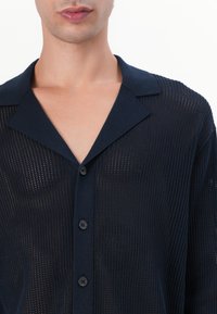 Navalinis mygtukų marškinėliai, pagaminti iš tinklelio audinio, su apykakle ir laisvu siluetu. Yra tekstūrinis raštas su matomais tinklelio tipo atvirais plotais.
