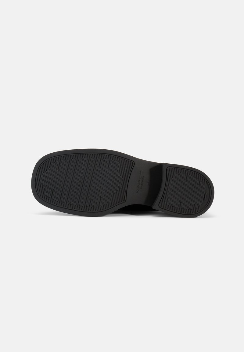 Suela de zapato negra con un patrón de agarre estriado y un pequeño logo en relieve visible cerca del arco sobre un fondo blanco.