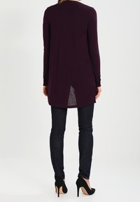 Kobieta ubrana w długą ciemnofioletową tunikę z rozcięciem z tyłu, ciemne skinny jeans i czarne szpilki, stoi tyłem.