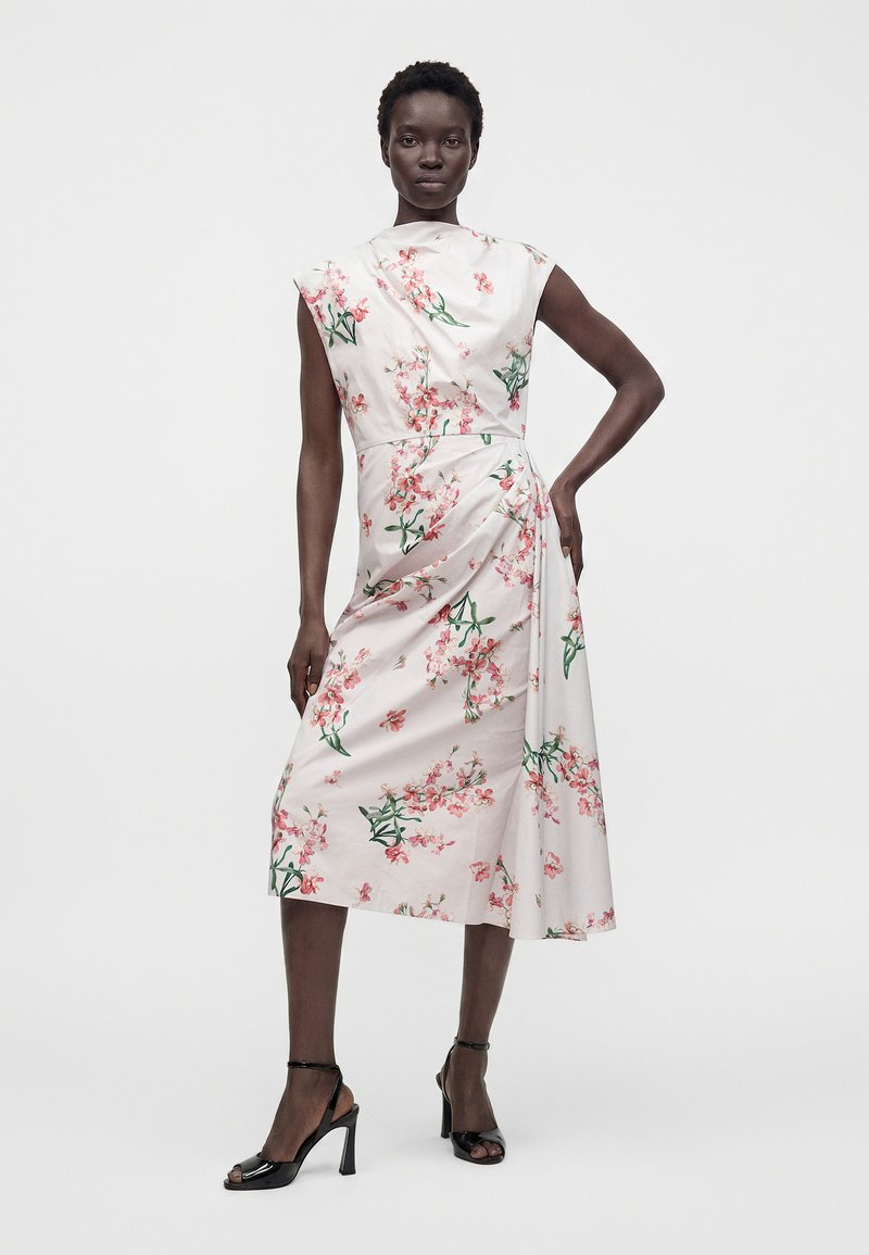 Woman wearing a sleeveless white midi dress with pink floral print and a draped detail, paired with black open-toe high-heeled sandals.