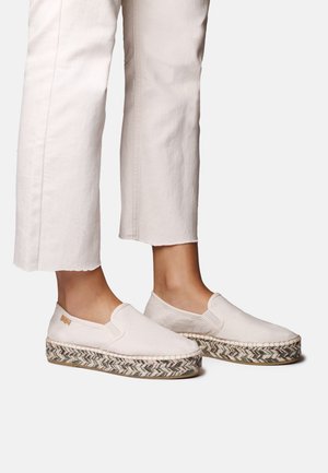Zapatos beige de lona tipo slip-on con una suela de esparto texturizada que presenta un patrón de espiga tejido y un pequeño detalle con el logo.