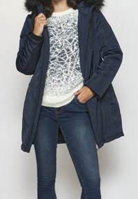 Parka acolchada azul marino con capucha de piel sintética, con cierre de cremallera y ajuste ligeramente holgado. Se lleva sobre un suéter ligero y unos jeans.