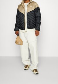 Chaqueta acolchada con la parte superior beige y la parte inferior negra, con cierre de cremallera y puños acanalados. Combinada con pantalones de color claro y zapatillas.