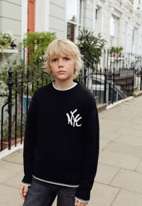 Černý pletený svetr s bílým logem NYC, žebrovaným límcem a manžetami, ve spojení s tmavě šedými džíny. V pozadí je obytná ulice.