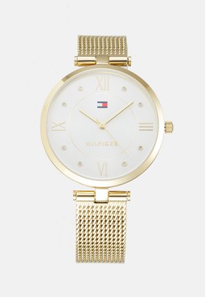 Montre avec un bracelet en maille dorée, un cadran rond, un cadran blanc, des chiffres romains, de petits joyaux comme marqueurs, et un détail de logo en rouge et bleu.