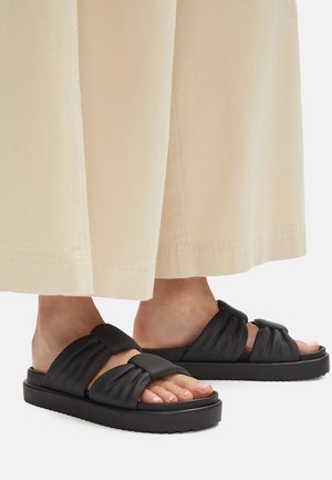 Czarne sandały z grubym, fakturowanym paskiem ozdobionym wiązaniem w formie supełka. Podeszwa jest gruba i z rowkami, zapewniając stabilną podstawę.