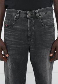 Svart denimjeans med en slitt tekstur, fem lommer og metallknapplukking. Har en liten rød merkeblubb på venstre lomme.