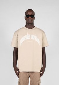 Beige bomull T-skjorte med korte ermer som har hvit grafisk tekst. Kombinert med cargo-shorts. Modell med mørke solbriller.