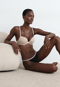 Beige gepolsterter BH mit dünnen Trägern und glatter Textur, kombiniert mit schwarzen Bikiniunterteilen mit einem kontrastierenden Bund, vor einem neutralen Hintergrund.