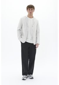 Helles beige Blazer mit zwei Knöpfen und Taschen, kombiniert mit einem weißen T-Shirt und schwarzen Hosen. Graue Sportschuhe vervollständigen das Outfit.