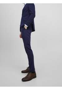 Námořnický modrý oblek s přizpůsobeným sakem a slim-fit kalhotami, doplněný o tmavě hnědé boty na šněrování, vyrobené z hladké kůže s jemnou texturou.