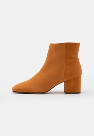 Lys brun ruskind ankle støvle med blokhæl og spids tå, vist fra siden på en ensfarvet baggrund.