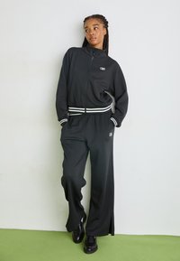 Completo sportivo nero con giacca zip-up e pantaloni a gamba larga. Caratterizzato da accenti a strisce bianche su polsini e cintura. Tessuto morbido.