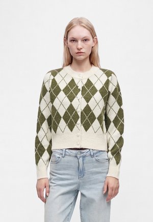 Ung kvinde med lige blondt hår iført en grøn og hvid argyle knapcardigan og lyseblå jeans, stående foran en ensfarvet baggrund.