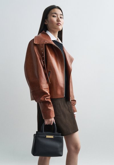 Brun læderjakke med forlængede ærmer, lynlås lukning og nitter, parret med en sort håndtaske og en brun ruskinds nederdel.