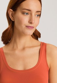 Top aderente senza maniche in tessuto color ruggine con una texture liscia. Presenta un ampio scollo e sottili spalline.
