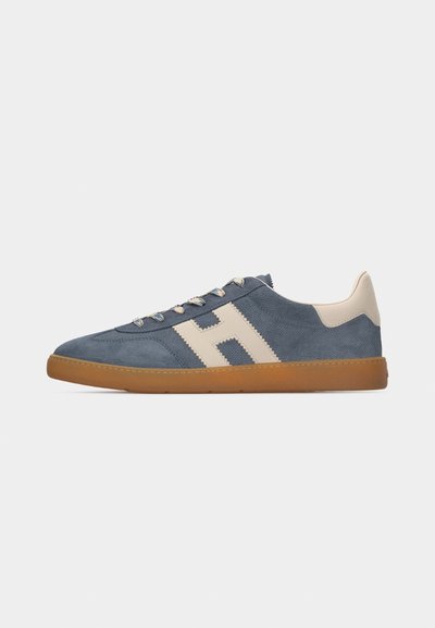 Baskets en daim bleu avec un logo "H" beige, un patch talon beige, une semelle en caoutchouc couleur gomme et des lacets à motifs sur un fond gris clair uni.
