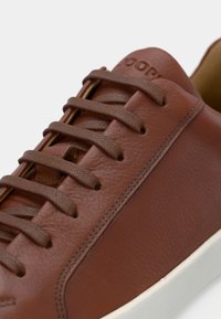 Brauner Ledersneaker mit strukturierter Oberfläche, passenden Schnürsenkeln und weißer Gummisohle. Logo ist an der Seite nahe der Oberseite sichtbar.