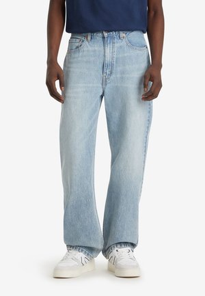 Homme portant un jean large bleu clair et des baskets blanches, debout devant un fond uni.